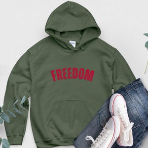Puede incluir: Una sudadera con capucha verde oliva con la palabra "FREEDOM" impresa en rojo en el pecho. La sudadera tiene un bolsillo delantero y una capucha con cord&oacute;n. Zapatillas blancas y vaqueros azules est&aacute;n junto a la sudadera.