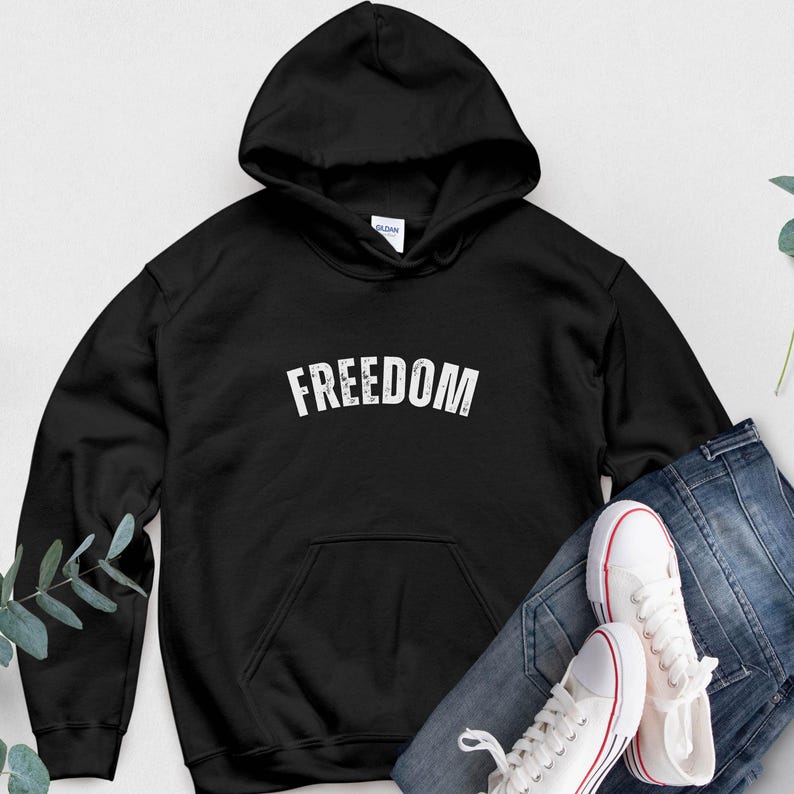 Puede incluir: Sudadera con capucha negra con la palabra "FREEDOM" en letras blancas. La sudadera tiene un bolsillo delantero y una capucha con cord&oacute;n. Un par de zapatillas blancas con detalles rojos y unos vaqueros azules est&aacute;n al lado.