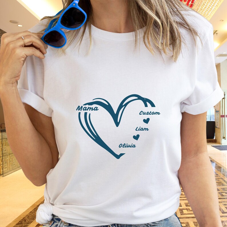 May include: White t-shirt featuring a teal heart design with the word 'Mama' on the left side of the heart. The right side of the heart has the word 'Custom' above a small heart, followed by 'Liam' and 'Olivia' below with small hearts. The shirt is plain white with rolled-up sleeves.