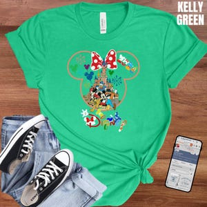 May include: Kelly green t-shirt featuring a Disney-themed graphic. The graphic includes a Minnie Mouse head outline with a red bow, a castle, and various Disney characters. The word "Disney" is also visible. The shirt is paired with jeans and black sneakers.