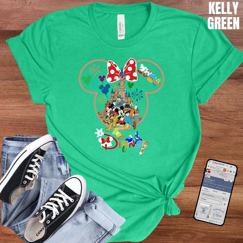 May include: Kelly green t-shirt featuring a Disney-themed graphic. The graphic includes a Minnie Mouse head outline with a red bow, a castle, and various Disney characters. The word "Disney" is also visible. The shirt is paired with jeans and black sneakers.