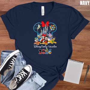 May include: Navy blue t-shirt featuring a colorful graphic of Disney characters, including Mickey and Minnie Mouse, in front of a castle. The text "Disney Family Vacation 2023" is also printed on the shirt. The shirt is paired with blue jeans and black sneakers.
