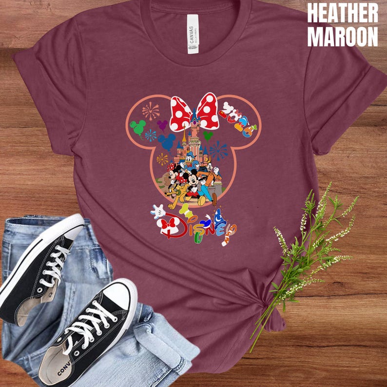 May include: Heather maroon t-shirt featuring a colorful Disney graphic. The design includes a Minnie Mouse bow, Disney characters, and the word "Disney". Black canvas sneakers and blue jeans are also in the image.