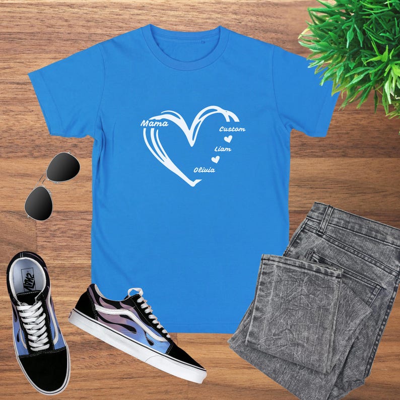 May include: A bright blue t-shirt featuring a white heart design with the words "Mama", "Custom", "Liam", and "Olivia". The shirt is paired with sunglasses, sneakers, and gray jeans on a wooden surface.