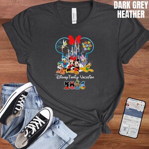 May include: Dark grey heather t-shirt featuring a colorful graphic of Disney characters in front of a castle, with the text "Disney Family Vacation 2023". The shirt is paired with blue jeans and black sneakers.