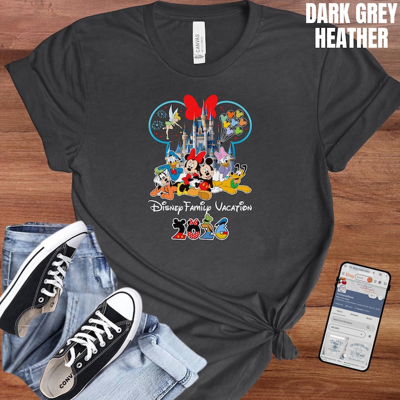 May include: Dark grey heather t-shirt featuring a colorful graphic of Disney characters in front of a castle, with the text "Disney Family Vacation 2023". The shirt is paired with blue jeans and black sneakers.