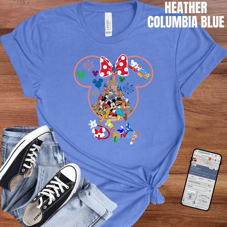 May include: Heather Columbia blue t-shirt featuring a colorful Disney graphic. The design includes Mickey and Minnie Mouse, a castle, and the word "Disney". The shirt is paired with black sneakers and blue jeans.