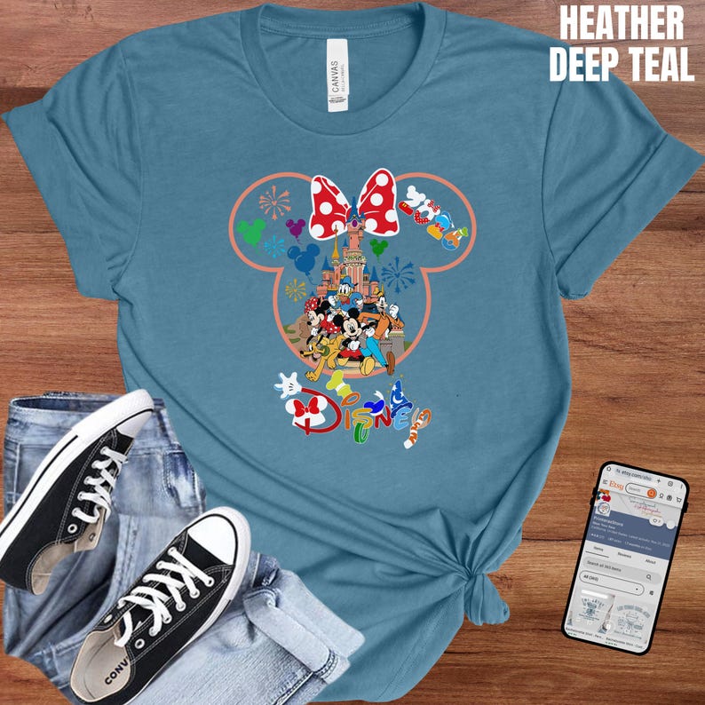 May include: Heather deep teal t-shirt featuring a Minnie Mouse design with a red and white polka dot bow, a castle, and various Disney characters. The shirt is paired with blue jeans and black Converse sneakers.