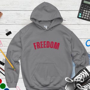 Puede incluir: Una sudadera con capucha gris con la palabra "FREEDOM" impresa en rojo en el pecho. La sudadera tiene un bolsillo delantero y una capucha con cord&oacute;n. La imagen est&aacute; sobre una superficie de vetas de madera clara.