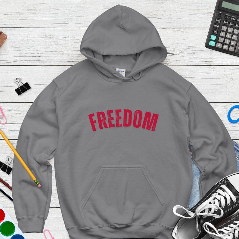 Puede incluir: Una sudadera con capucha gris con la palabra "FREEDOM" impresa en rojo en el pecho. La sudadera tiene un bolsillo delantero y una capucha con cord&oacute;n. La imagen est&aacute; sobre una superficie de vetas de madera clara.