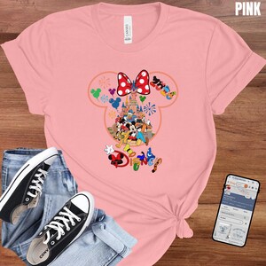 May include: Pink t-shirt featuring a colorful Disney graphic. The design includes Mickey and Minnie Mouse, a castle, and the word "Disney". The shirt is paired with black Converse sneakers and blue jeans.