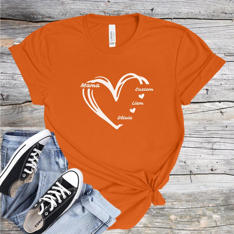 May include: Orange t-shirt with a white heart design. The heart contains the word "Mama" and the names "Liam" and "Olivia", with the word "Custom" above them. The shirt is paired with blue jeans and black sneakers.