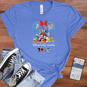 May include: A light blue t-shirt featuring a colorful graphic of Disney characters, including Mickey and Minnie Mouse, in front of a castle. The text "Disney Family Vacation 2023" is printed below the characters.