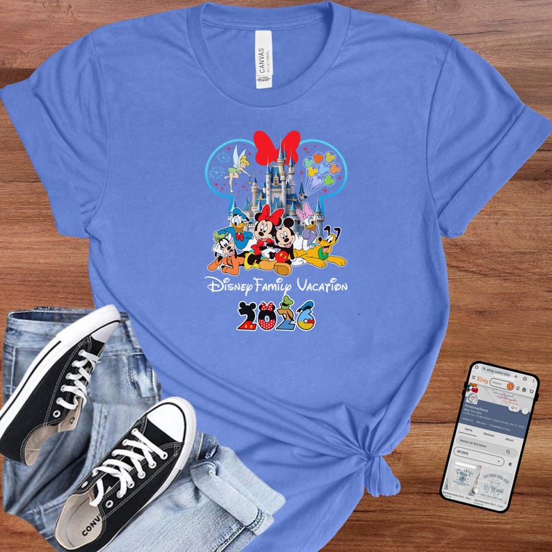 May include: A light blue t-shirt featuring a colorful graphic of Disney characters, including Mickey and Minnie Mouse, in front of a castle. The text "Disney Family Vacation 2023" is printed below the characters.