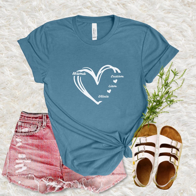May include: A teal t-shirt with a white heart design. The heart contains the word "Mama" and the names "Liam" and "Olivia". The word "Custom" is also in the heart design. The shirt is paired with pink shorts and sandals.