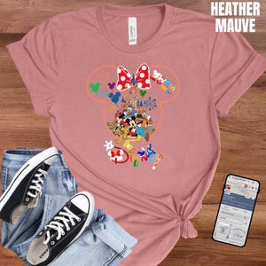 May include: Heather mauve t-shirt featuring a colorful Disney graphic. The graphic includes a castle, Mickey Mouse, Minnie Mouse, and other Disney characters. The word "Disney" is also visible. The shirt is paired with jeans and black sneakers.