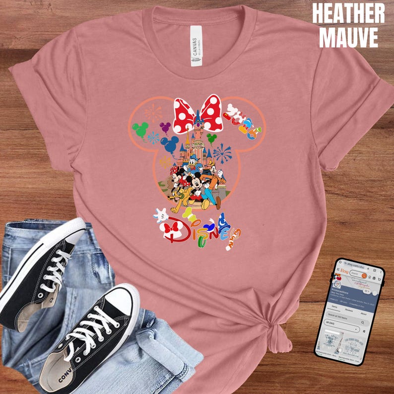 May include: Heather mauve t-shirt featuring a colorful Disney graphic. The graphic includes a castle, Mickey Mouse, Minnie Mouse, and other Disney characters. The word "Disney" is also visible. The shirt is paired with jeans and black sneakers.