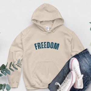 Puede incluir: Una sudadera con capucha color crema con la palabra "FREEDOM" impresa en letras turquesa desgastadas en el pecho. La sudadera tiene un bolsillo delantero y una capucha. Un par de zapatillas de lona blancas con detalles rojos y unos vaqueros azules est&aacute;n junto a la sudadera.