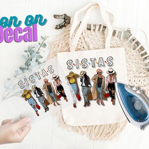 May include: Iron-on decal featuring a group of five women wearing hats and stylish outfits. The decal says "Sistas" in colorful letters.