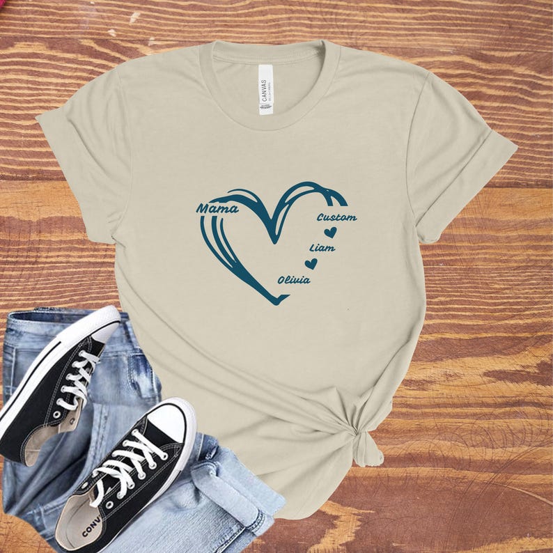 May include: A beige t-shirt with a navy blue heart design. The heart contains the word "Mama" and the names "Liam" and "Olivia". The word "Custom" is also included. Black and white sneakers and blue jeans are in the image.