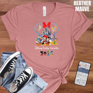May include: Heather mauve t-shirt featuring a colorful Disney Family Vacation design with Mickey and Minnie Mouse, Donald Duck, and other characters. The design includes a castle and the year 2026. Black canvas sneakers and blue jeans are also in the image.
