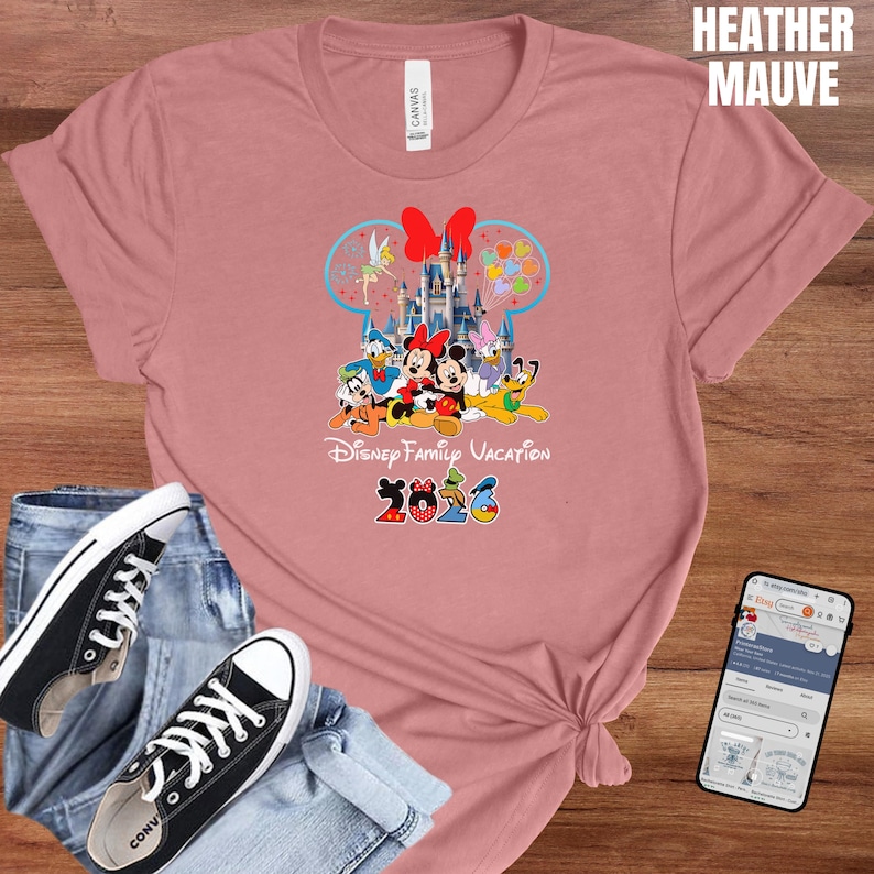 May include: Heather mauve t-shirt featuring a colorful Disney Family Vacation design with Mickey and Minnie Mouse, Donald Duck, and other characters. The design includes a castle and the year 2026. Black canvas sneakers and blue jeans are also in the image.