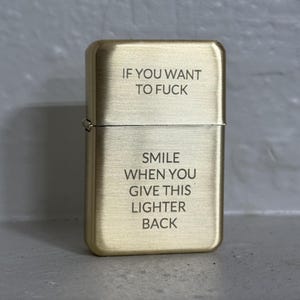 May include: A brass-colored metal lighter with engraved text. The text reads "IF YOU WANT TO FUCK SMILE WHEN YOU GIVE THIS LIGHTER BACK."