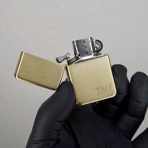 May include: A brass-colored lighter with the lid open, revealing the interior. The lighter has a rectangular shape with a brushed metal finish. The initials "TMF" are engraved on the front. The lighter is held by a person wearing a black glove.