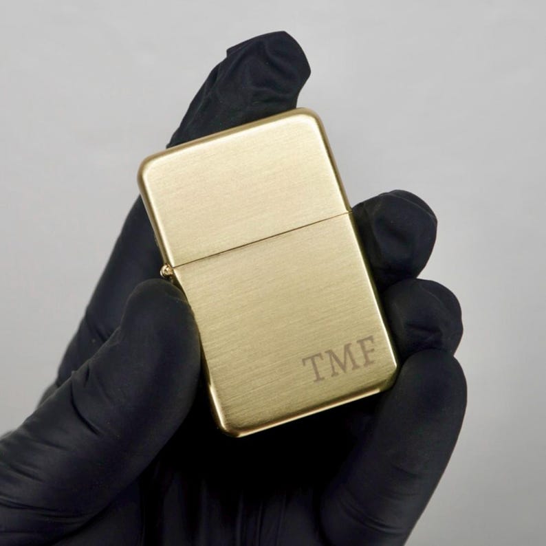 May include: A gold-colored lighter with a brushed metal finish. The lighter has the initials "TMF" engraved on the front. The lighter is held in a black gloved hand against a neutral background.