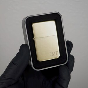 May include: A gold-colored lighter with the initials "TMF" engraved on the front, presented in a silver metal case with a black foam insert. The lighter is held by a gloved hand.