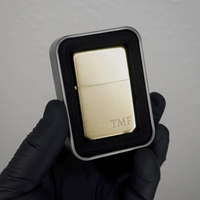 May include: A gold-colored lighter with the initials "TMF" engraved on the front, presented in a silver metal case with a black foam insert. The lighter is held by a gloved hand.
