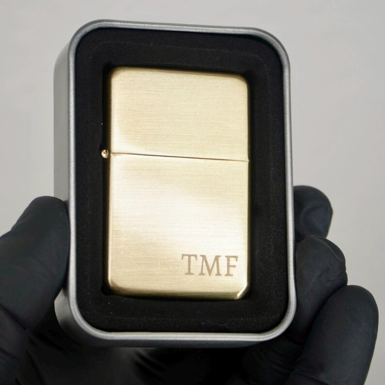 May include: A gold-colored lighter with the initials "TMF" engraved on the front. The lighter is in a silver metal case with a black foam insert. The lighter is rectangular and has a brushed metal finish.