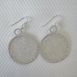 Peut inclure: Une paire de boucles d'oreilles ajourées argentées. Chaque boucle d'oreille présente un motif circulaire avec un motif délicat et tissé. Les boucles d'oreilles ont une fermeture à crochet pour oreilles percées. Elles sont sur une surface blanche texturée.