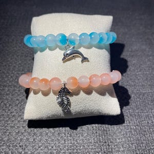 Könnte beinhalten: Zwei Armbänder mit Perlen. Eines ist hellblau mit einem silbernen Delphin-Anhänger, das andere ist pfirsichfarben mit einem silbernen Seepferdchen-Anhänger. Die Armbänder werden auf einem weißen Kissen vor grauem Hintergrund präsentiert.