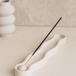 May include: A white ceramic incense holder with a wavy design, holding a black incense stick. A white, stacked vase is in the background. The holder has a small, round base to catch ash.