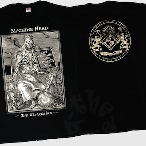 Puede incluir: Camiseta negra con el nombre de la banda "MACHINE HEAD" y una ilustración detallada en blanco y negro de un esqueleto sosteniendo un letrero. El texto "The Blackening" está debajo. La parte trasera tiene un emblema dorado.