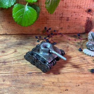 May include: A miniature metal tank sculpture, crafted from metal chain links, a large nut, and a bolt. The tank has a silver finish and is positioned on a wooden surface. Green leaves and berries are visible in the background.