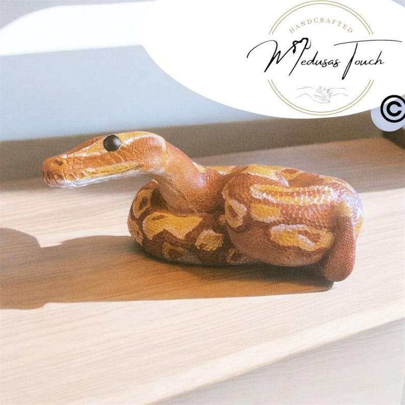 May include: A handcrafted, realistic sculpture of a brown and yellow snake with a white belly. The snake is curled up in a resting position with its head raised. The sculpture is made by Medusas Touch.