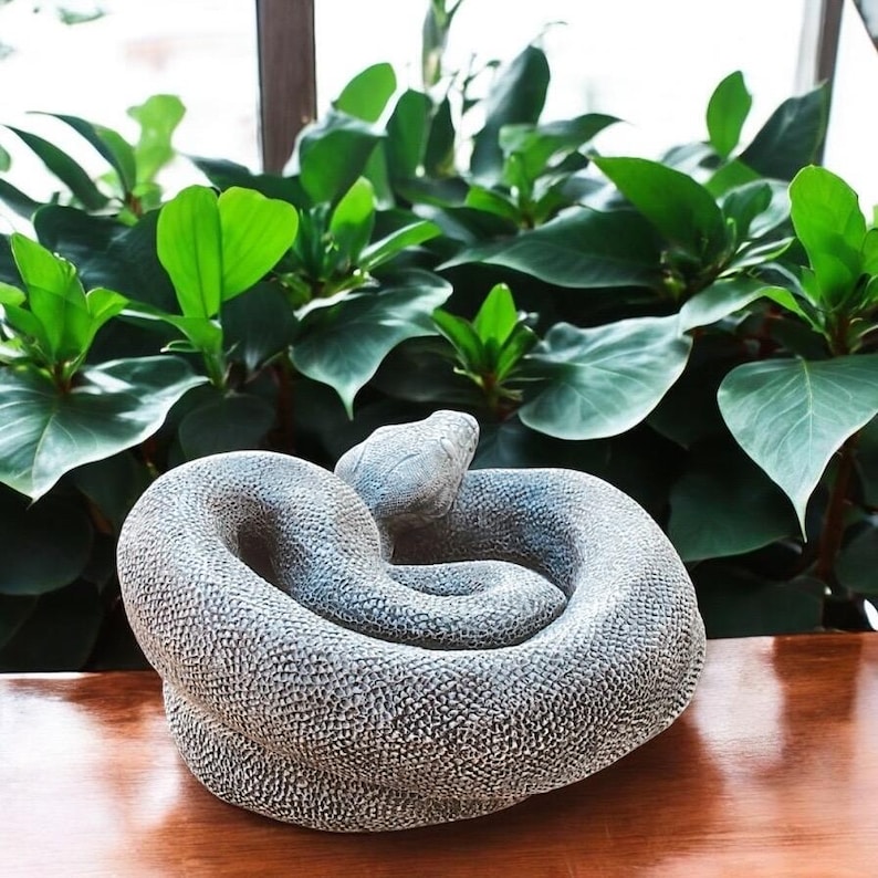 以下が含まれることがあります： テクスチャ加工された表面を持つ、グレーの巻き付けられたヘビの彫刻。彫刻は石のような素材で作られています。