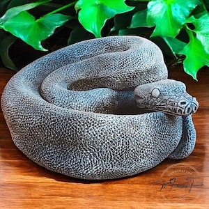 以下が含まれることがあります： 灰色のコンクリート製の蛇のプランターで、円形に巻かれ、頭が体に置かれています。プランターは、蛇の鱗のような質感のある表面をしています。プランターは手作りで、底に「The Midas Touch」という文字が書かれています。