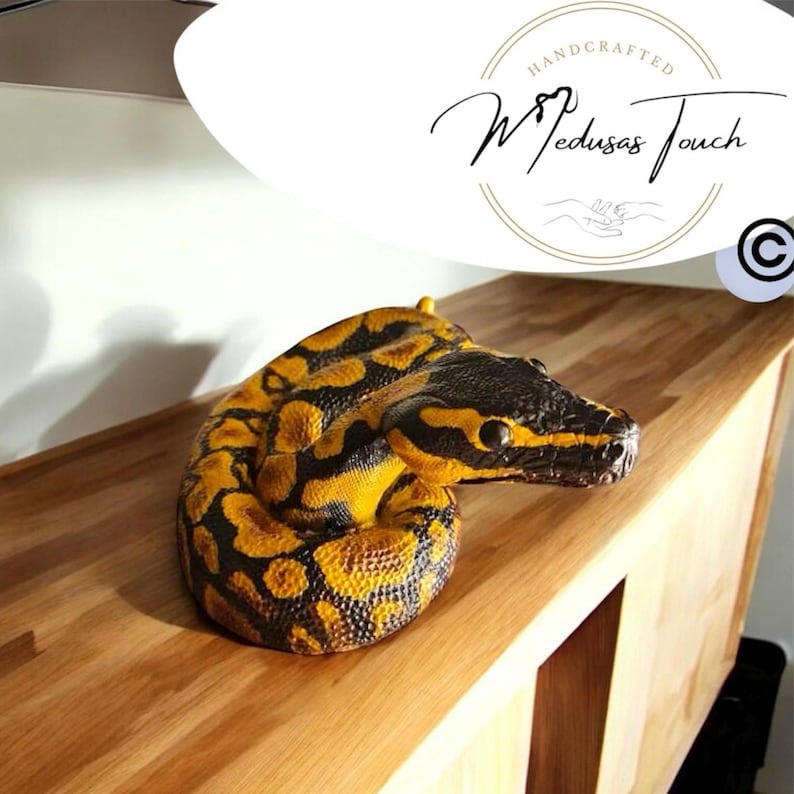 May include: A yellow and black snake with a patterned design is coiled on a wooden surface. The snake is a handcrafted item and the text "Medusas Touch" is visible in the background.