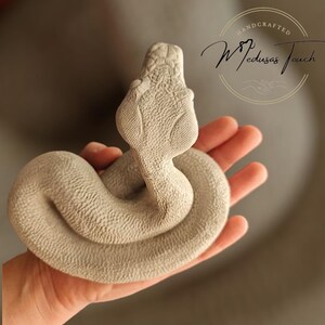 May include: A handcrafted, white ceramic snake sculpture. The snake is coiled in a relaxed pose with its head raised. The sculpture is detailed with realistic scales and a textured surface.