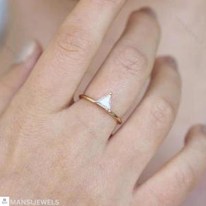 May include: A gold ring with a triangular, white gemstone. The ring has a slender band and a minimalist design. The gemstone is set in a delicate prong setting. The ring is displayed on a finger.