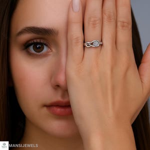以下が含まれることがあります： 結び目のデザインと小さな透明な石のクラスターが付いたシルバーリング。 リングは人の指にあり、手で顔の一部を覆っています。 リングが画像の中心です。