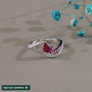 May include: A silver ring with a large, red, kite-shaped gemstone at its center. The ring features a band of small, clear stones and a curved section with smaller red stones. The ring is set against a neutral background.