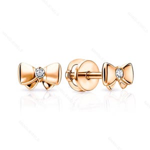May include: Rose gold bow stud earrings. Each earring has a bow design with a small, round, clear gemstone in the centre. The earrings are displayed on a white background, highlighting their polished finish.