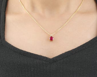 May include: A gold necklace featuring a teardrop-shaped red gemstone pendant. The pendant is adorned with small, clear stones. The necklace is worn against a black top.