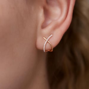 以下が含まれることがあります： ローズゴールドのX字型スタッドピアス。片側に小さなクリアクリスタルが並んでいます。ピアスは耳に装着されています。モダンでミニマルなデザインです。