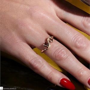 May include: A gold ring with two oval-shaped, reddish-brown gemstones. The ring has a unique, flowing design, with the gemstones set in a wave-like pattern. The ring is shown on a hand with red nail polish.
