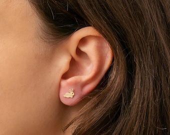 May include: A gold bunny stud earring is displayed on an ear. The earring is a small, minimalist design, featuring a detailed outline of a rabbit. The earring is a delicate piece of jewellery.
